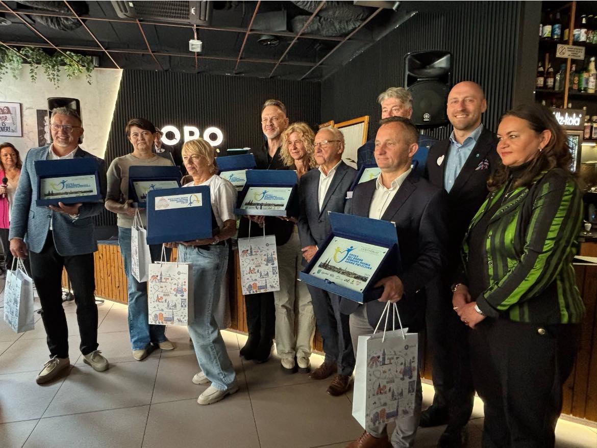 Zdjęcie grupowe, w środku stoi Pan Mateusz Szpryngiel, Koordynator ds. Dostępności UM Opola, wokół niego uśmiechnięci seniorzy w jednakowych koszulkach