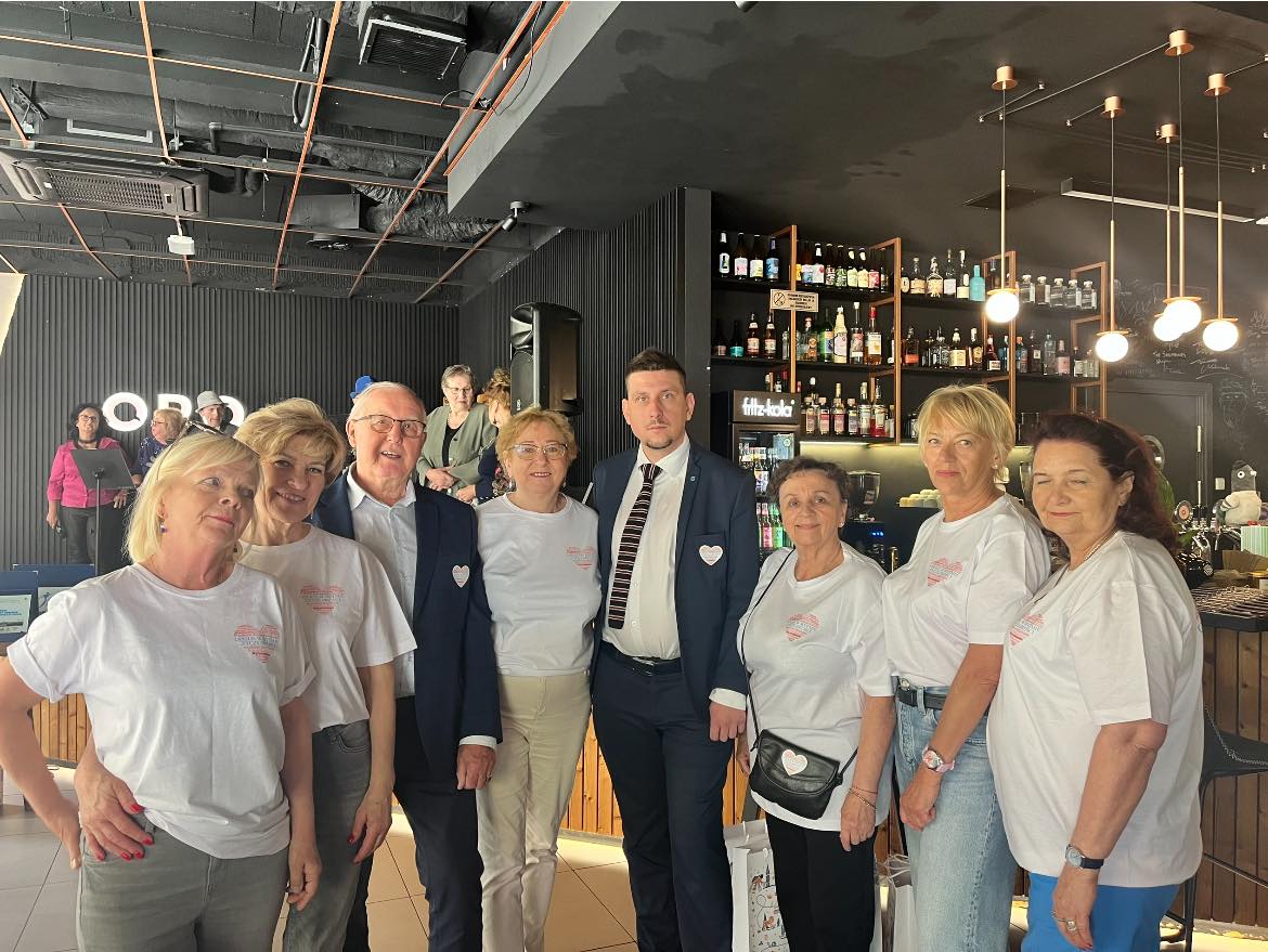 Zdjęcie grupowe, w środku stoi Pan Mateusz Szpryngiel, Koordynator ds. Dostępności UM Opola, wokół niego uśmiechnięci seniorzy w jednakowych koszulkach