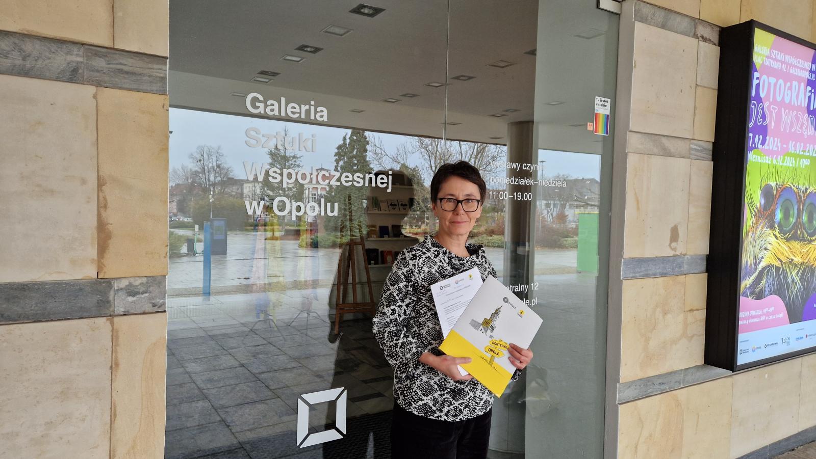 Dyrektor Galerii Sztuki Współczesnej w Opolu, Pani Joanna Filipczyk trzymająca w rękach porozumienie o współpracy i teczkę Dostępne Opole. W tle wejście główne do galerii