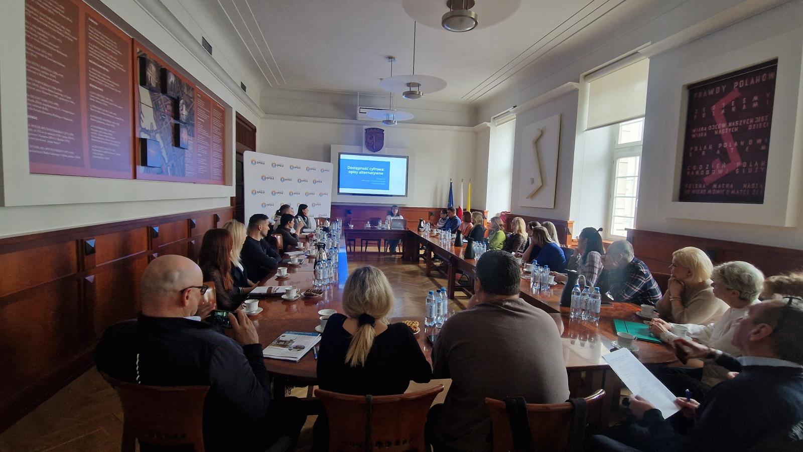 Grupa uczestników siedzących przy dużym okrągłym stole w sali Ratusza. Na wprost ekran z prezentacją, po lewej stronie ścianka z logotypami Przebojowe Opole.