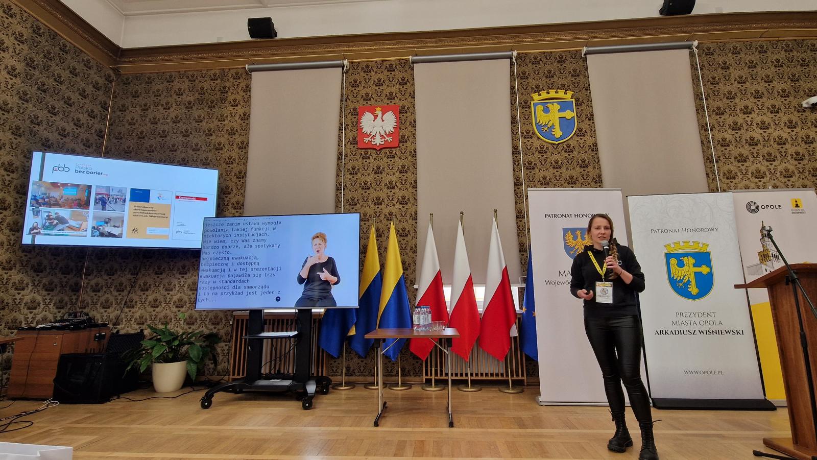 Sala w opolskim Ratuszu. Przy mównicy stoi Pani Joanna Piwowońska, Kobieta ubrana w sukienkę i buty na obcasie, ciemne, proste włosy do ramion