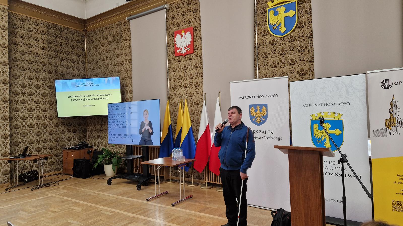 Sala w opolskim Ratuszu. Do mikrofonu mówi Pan Roman Roczeń. Mężczyzna w średnim wieku niewidomy z białą laską. Obok niego stoi plecak przy mównicy. Po lewej stronie ekrany z prezentacją i tłumaczem języka migowego, bo drugiej roll upy z patronatami honorowymi. 
