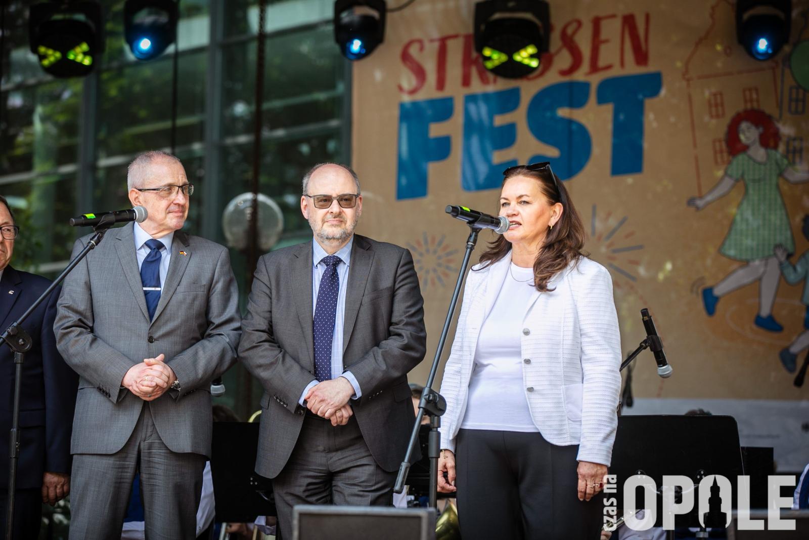 biała kobieta przemawia do mikrofony, obok dwóch białych mężczyzn patrzy w jej kierunku na tle sceny