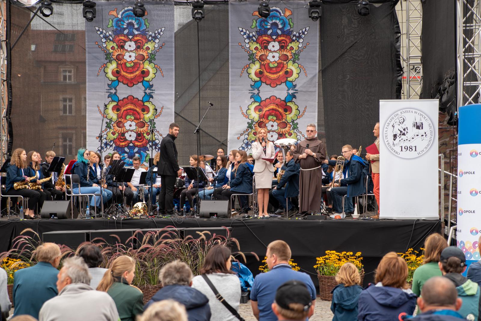 zdjęcie przedstawiające franciszkanina na scenie w towarzystwie orkiestry i prowadzącej wydarzenie kobiety