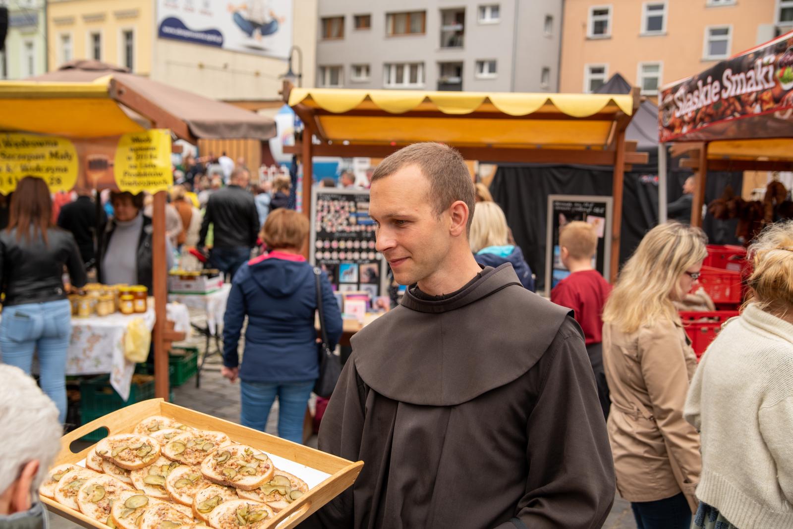 Franciszkanin niosący tacę z kanapkami, w tle stoiska jarmarczne