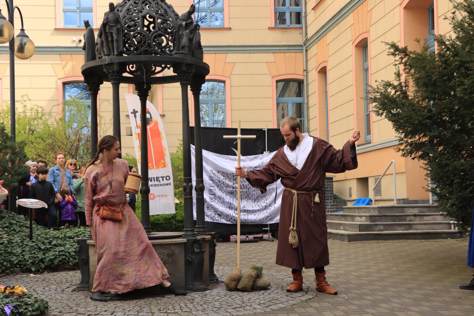 biała kobieta i biały mężczyzna, jako aktorzy, występują obok studni na Wzgórzu Uniwersyteckim podczas wydarzenia pt. Święto Wojciechowe