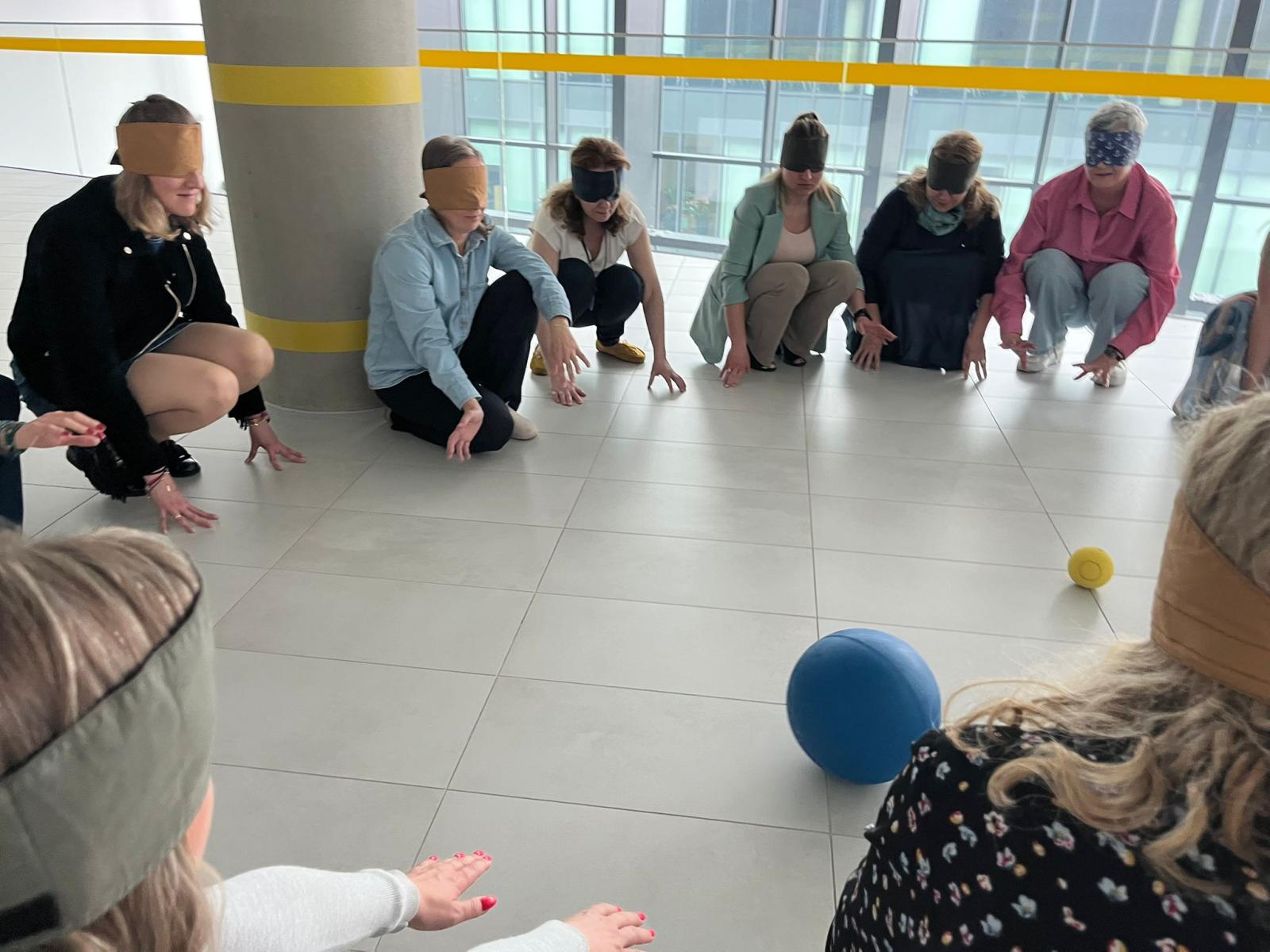 Grupa ludzi kuca w kółku. Wszyscy mają opaski na oczach. Na środku dwie piłki, jedna mała i jedna duża. 