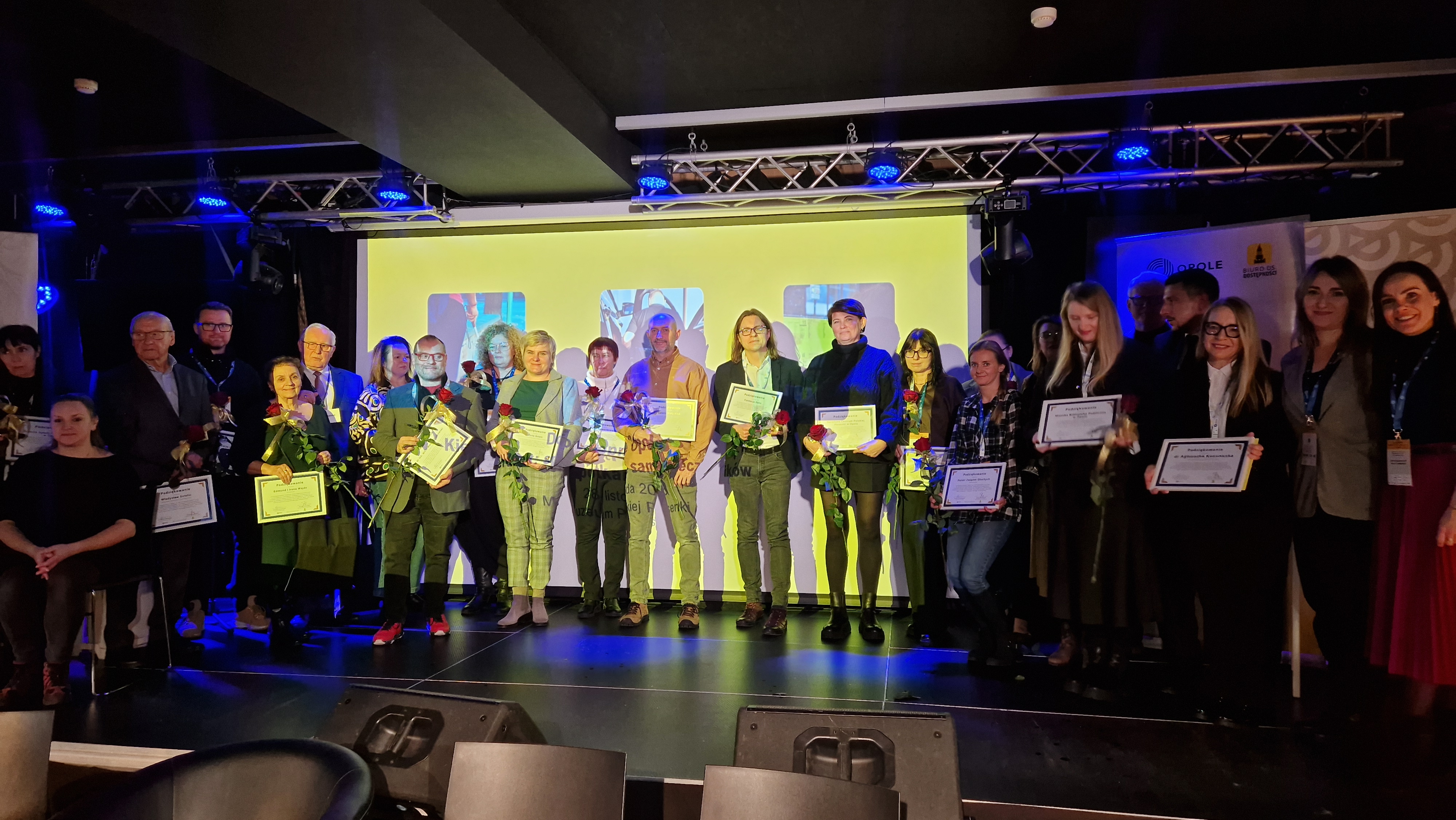 Zdjęcie grupowe uczetsników na scenie z dyplomami i różami.