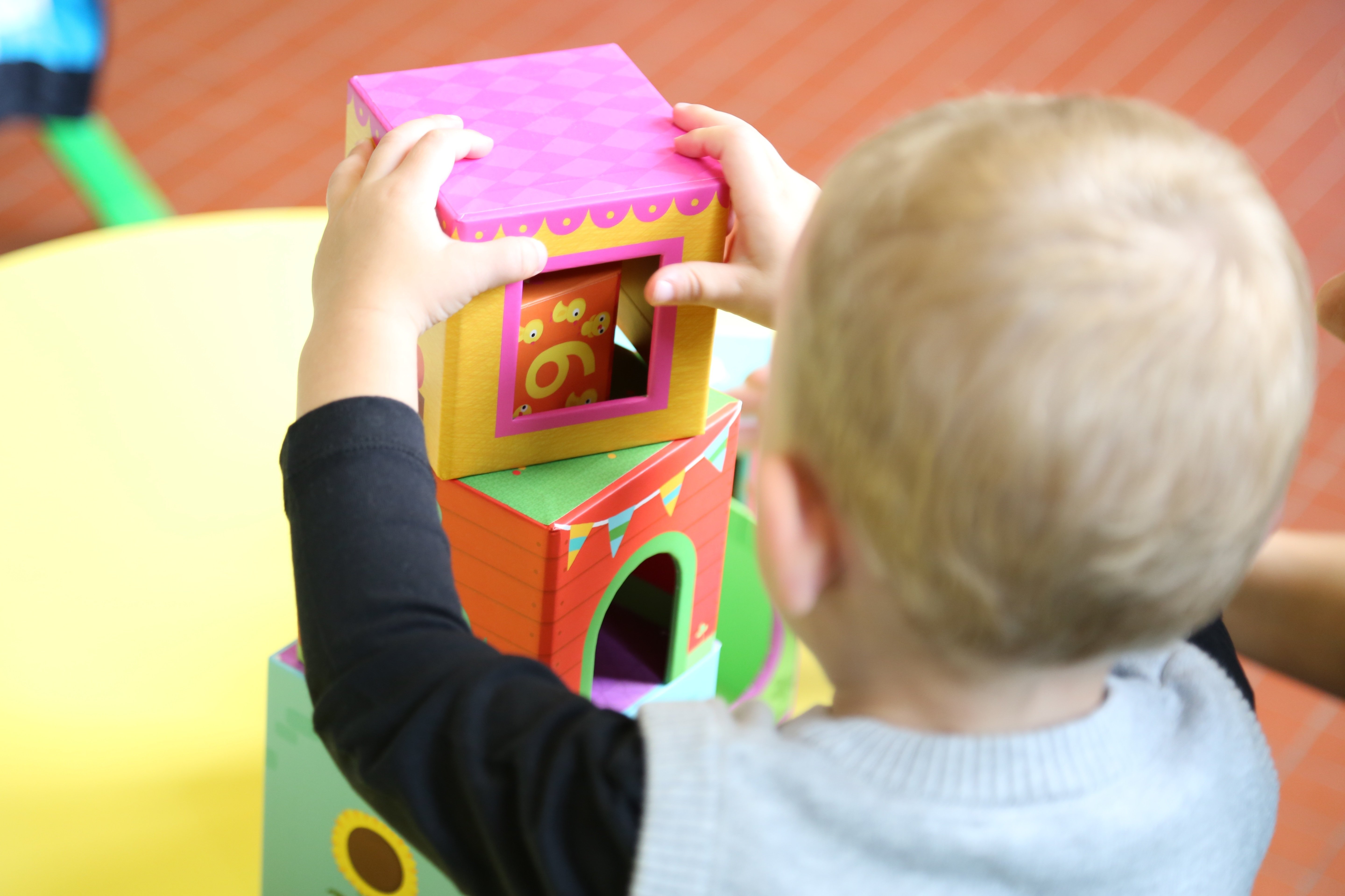 Małe dziecko w wieku do lat 3, widziane od tyłu, które oburącz układa wieżę z dużych, kolorowych kostek na stole. 