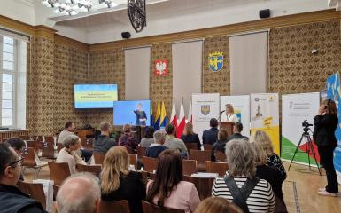 Sala w opolskim Ratuszu. Uczestnicy wydarzenia siedzą na krzesłach w układzie kinowym. Z przodu dwa telewizory, jeden z prezentacją, drugi z tłumaczem języka migowego. Przy mównicy stoi kobieta prowadząca Opolskie Forum Koordynatorów