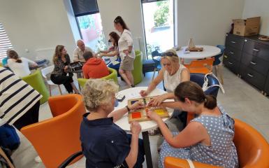 grupa ludzi siedzi przy okrągłych stołach i grają w różne gry planszowe, na pierwszym planie trzy kobiety grają w chińczyka dedykowanego dla osób niewidomych