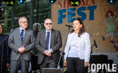biała kobieta przemawia do mikrofony, obok dwóch białych mężczyzn patrzy w jej kierunku na tle sceny