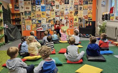 zdjęcie przedstawia dzieci siedzące na podłodze w bibliotece, ososba kobieta prowadzi zajęcia czytelnicze