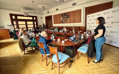 Sala Ratusza, uczestnicy szkolenia z dostępności cyfrowej siedzą przy stole i patrzą na prezentację i kobietę prowadzącą spotkanie.