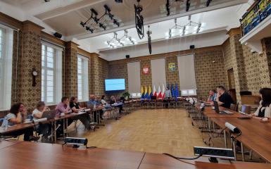 Zdjęcie pokazujące całą salę 312 w Ratuszu, stoły w układzie podkowy. Grupa uczestników szkolenia siedzi przy stole z laptopami. w Tle slajd z prezentacji