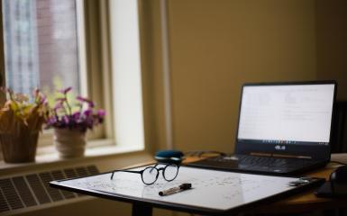 okulary w czarnych oprawkach, obok nich mazak biało-czarny, połozone na blacie biurka, w tle otwarty laptop z białym ekranem, na parapecie stoja dwie doniczki z kwatami domowymi