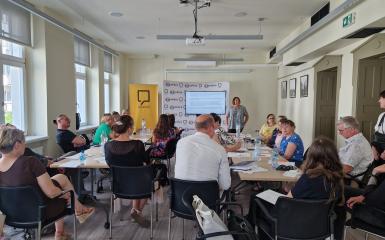 Grupa ludzi siedzących przy stołach, słuchający prowadzącą szkolenie. W tle slajd na monitorze.