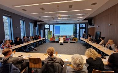 sala konferencyjna Miejskiej Biblioteki Publicznej w Opolu, grupa ludzi siedzących przy stołach, duży slajd prezentacji w tle. 