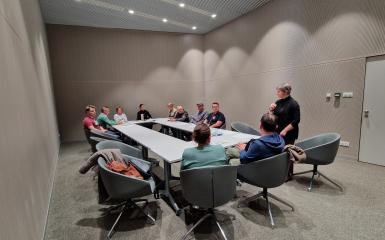 Uczestnicy szkolenia w salce konferencyjnej siedzą wokół podłużnego stołu. Prowadząca szkolenie stoi z boku. 