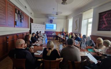 Grupa uczestników siedzących przy dużym okrągłym stole w sali Ratusza. Na wprost ekran z prezentacją, po lewej stronie ścianka z logotypami Przebojowe Opole. 