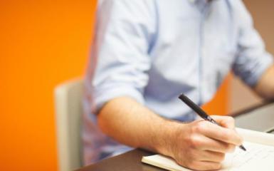 Osoba siedząca przy stole i pisząca na kartce, pomarańczowe tło