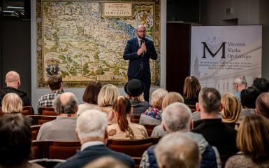 otwarcie wystawy, siedzący ludzie i przemawiający marszałek