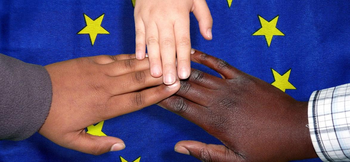 Niebieska flaga żółtymi gwiazdami położna na płasko. Na niej ułożone 3 ręce w kształt gwiazdy.