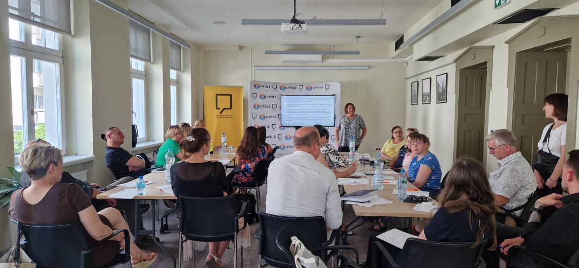 Grupa ludzi siedzących przy stołach, słuchający prowadzącą szkolenie. W tle slajd na monitorze.