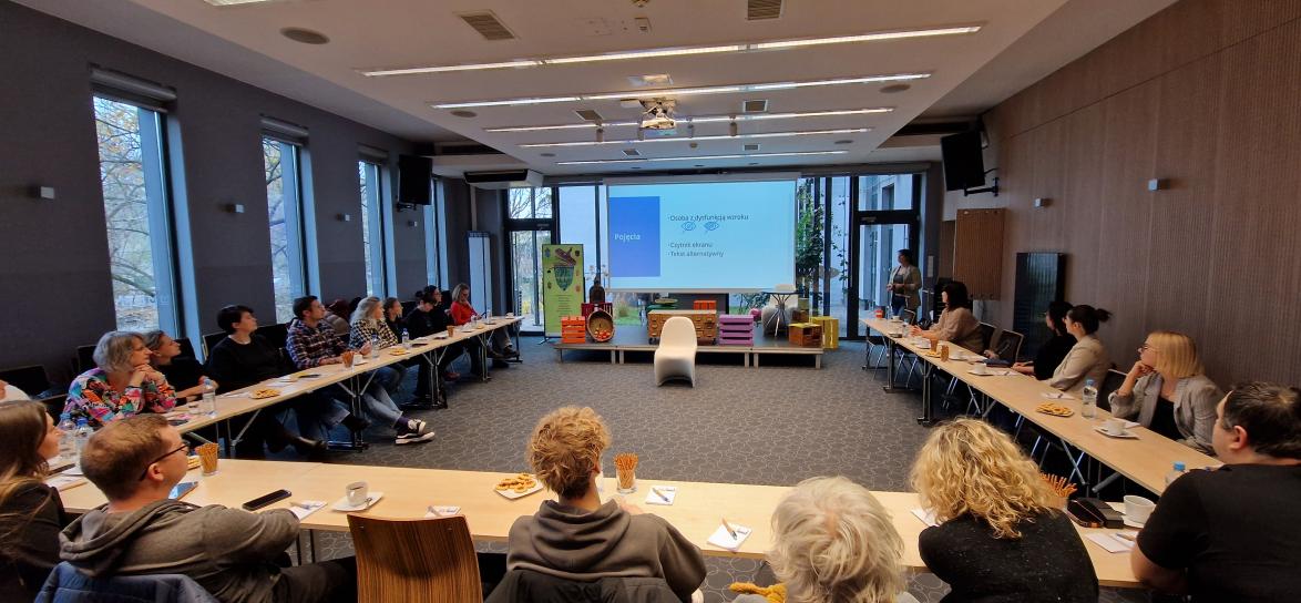 sala konferencyjna Miejskiej Biblioteki Publicznej w Opolu, grupa ludzi siedzących przy stołach, duży slajd prezentacji w tle. 