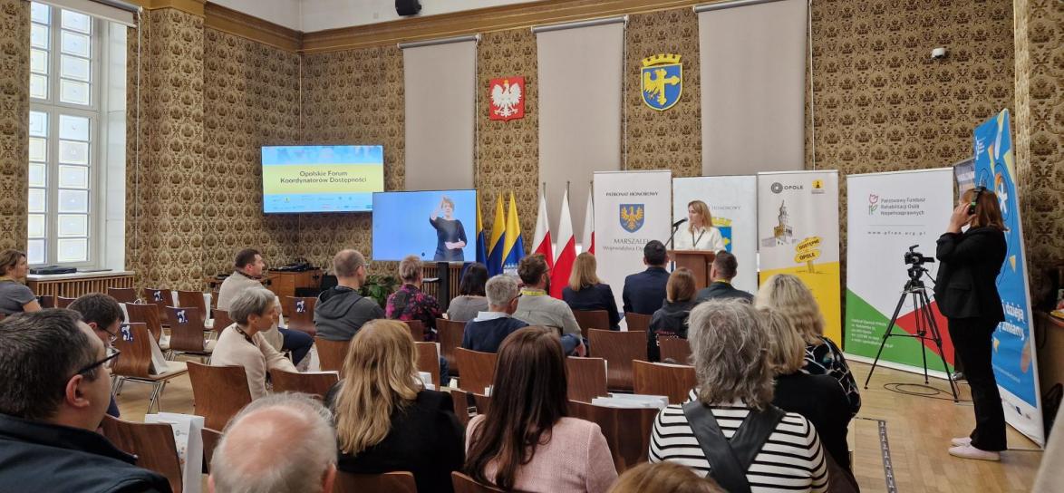 Sala w opolskim Ratuszu. Uczestnicy wydarzenia siedzą na krzesłach w układzie kinowym. Z przodu dwa telewizory, jeden z prezentacją, drugi z tłumaczem języka migowego. Przy mównicy stoi kobieta prowadząca Opolskie Forum Koordynatorów