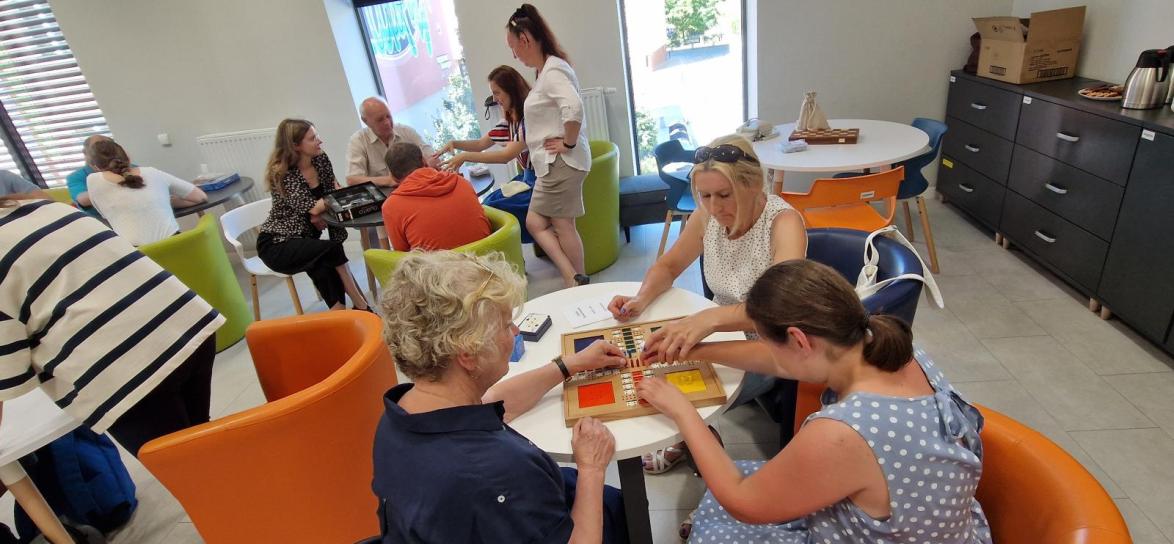 grupa ludzi siedzi przy okrągłych stołach i grają w różne gry planszowe, na pierwszym planie trzy kobiety grają w chińczyka dedykowanego dla osób niewidomych