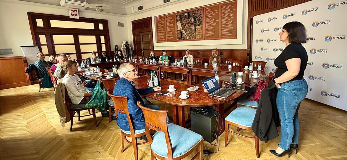 Sala Ratusza, uczestnicy szkolenia z dostępności cyfrowej siedzą przy stole i patrzą na prezentację i kobietę prowadzącą spotkanie.