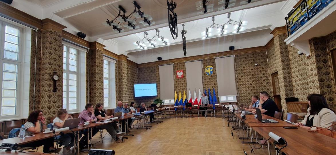 Zdjęcie pokazujące całą salę 312 w Ratuszu, stoły w układzie podkowy. Grupa uczestników szkolenia siedzi przy stole z laptopami. w Tle slajd z prezentacji