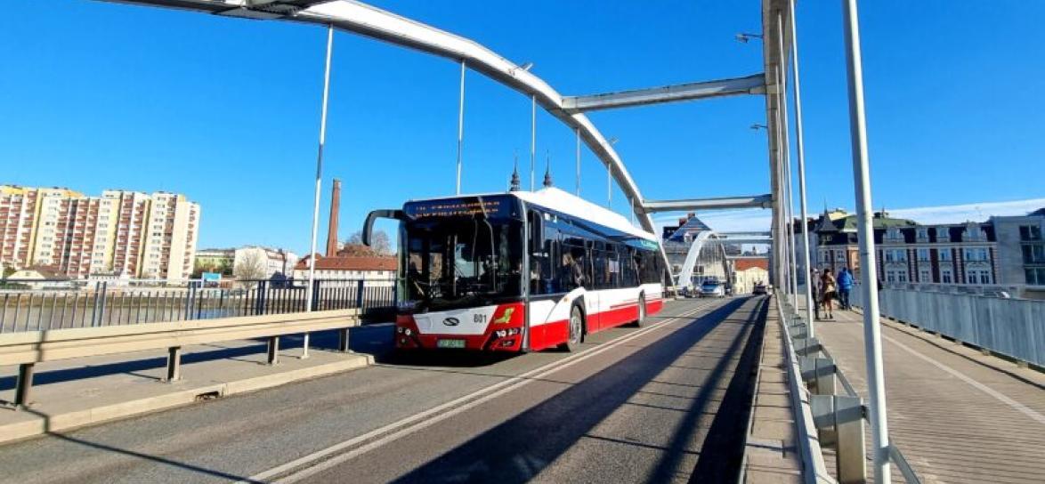 Most na rzeką. Na moście autobus. W tle wysokie bloki z wielkiej płyty. Pogodny dzień.