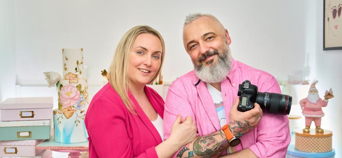 Para. Właściciele restauracji. Urbrani na różowo. Ona trzyma go pod ramię. On trzyma w dłoni aparat fotograficzny
