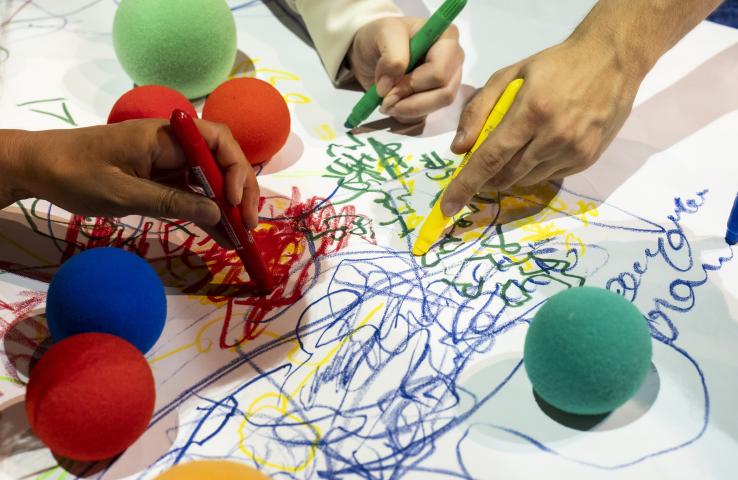 Zdjęcie przedstawia wiele rak rysujących po kartce papieru i kolorowe kuleczki