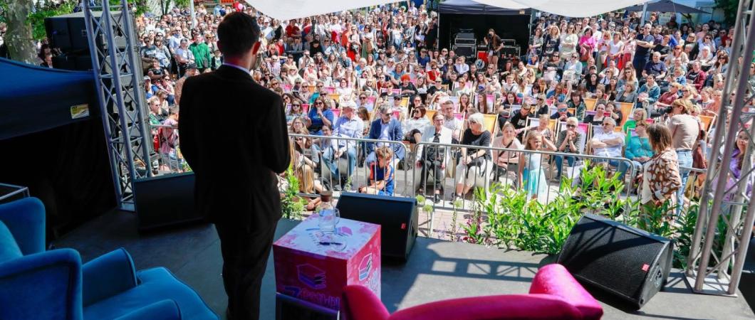 Biały mężczyzna w czarnym garniturze na scenie, stoi tyłem do obiektywu, na tle uczestników patrzących w jego kierunku