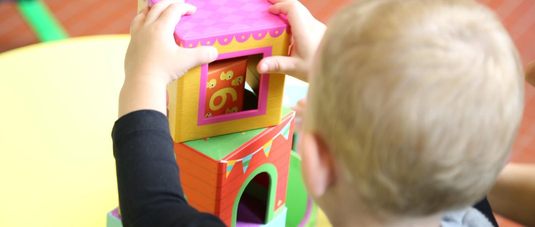 Małe dziecko w wieku do lat 3, widziane od tyłu, które oburącz układa wieżę z dużych, kolorowych kostek na stole. 